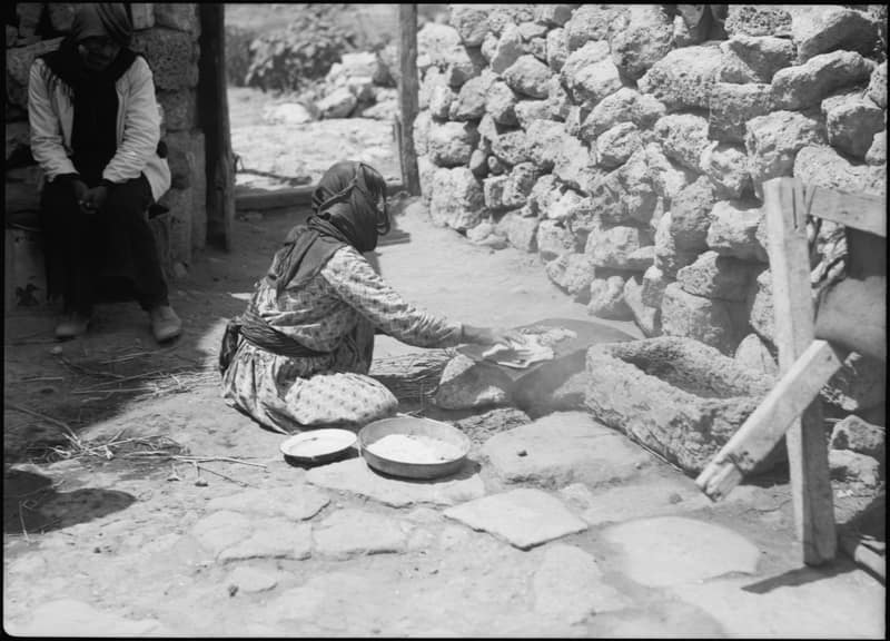 A woman kneels on packed earth and reaches forward to lay a thin sheet of dough over a low domed griddle set on a fire. Two metal basins of dough stand beside her knee. Behind her a rough wall is built of reused stone blocks and a man in a head-cloth sits watching from a doorway.