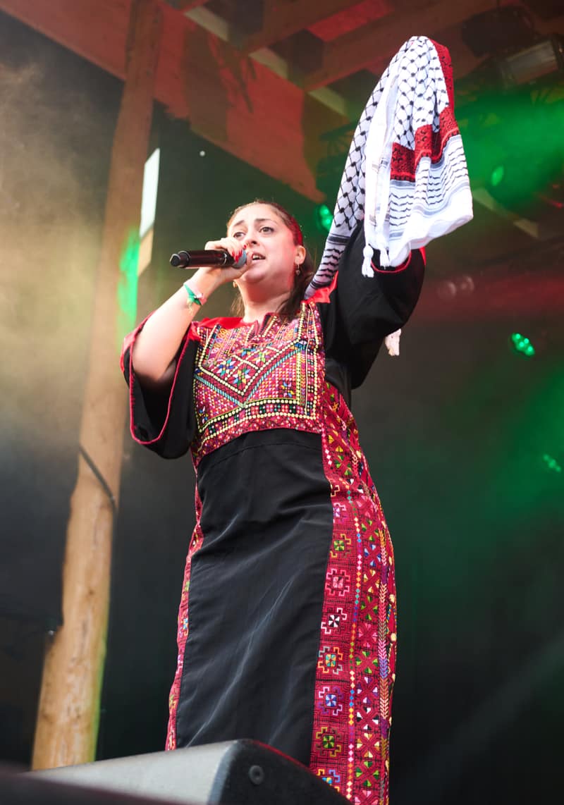 A singer photographed from below on a stage under green lights, holding a microphone to her mouth and raising a black-and-white keffiyeh above her head. She wears a black thobe with a densely cross-stitched chest panel and a wide embroidered panel running the full length of the skirt.