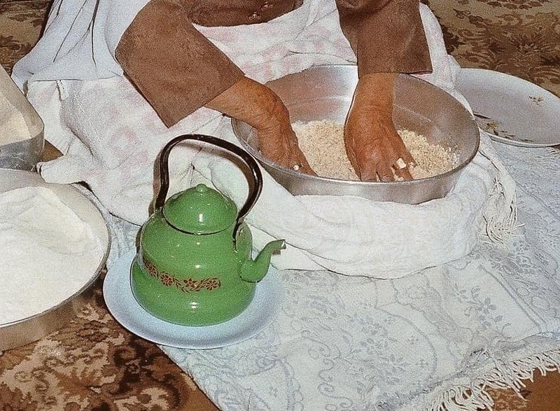 A close colour photograph of two hands working a mound of pale grain in a metal bowl set on a white cloth. The woman’s brown sleeves are pushed back; a green enamel teapot on a saucer and two trays of flour sit around her on a patterned carpet.