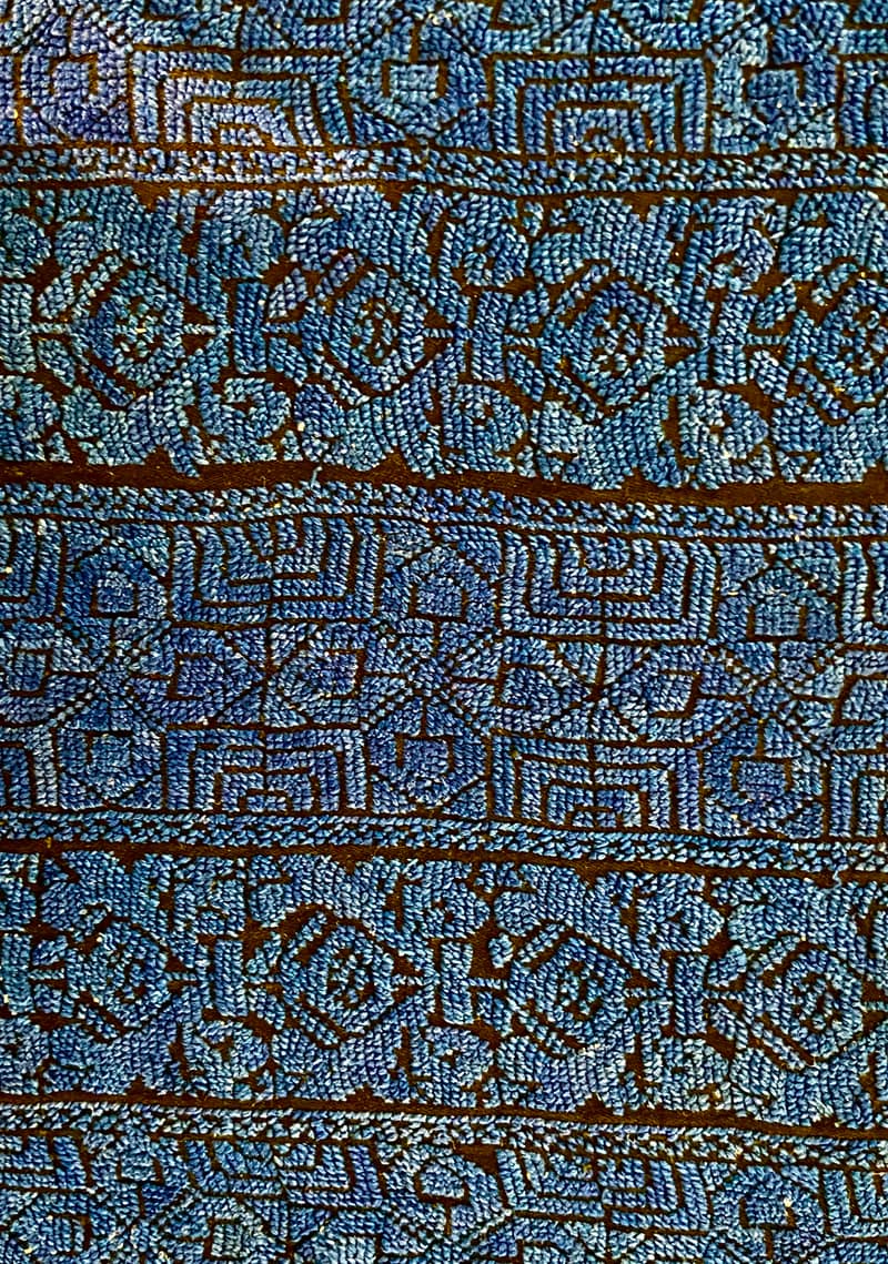 Dense bands of blue cross-stitch worked in geometric blocks across a dark brown ground, the pale blue thread almost covering the cloth.