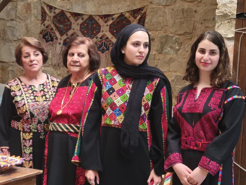 Four women of different ages stand side by side in front of a stone wall hung with a patterned kilim, each in a black thobe embroidered differently: one with a magenta chest panel and rose-worked cuffs, one with a chest panel of multicoloured diamonds, one with a crimson couched panel under a long gold chain.