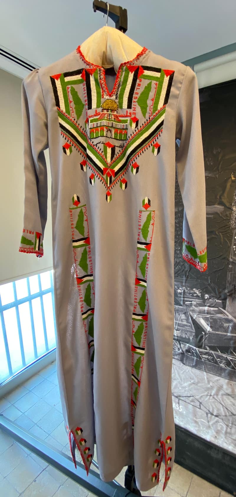 A long grey dress on a hanger behind museum glass. Cross-stitch in red, green, black and white forms a wide chest yoke around an embroidered Dome of the Rock with a gold cupola; the outline of Palestine in green and the word PALESTINE in red capitals repeat down two vertical panels of the skirt and around both cuffs.