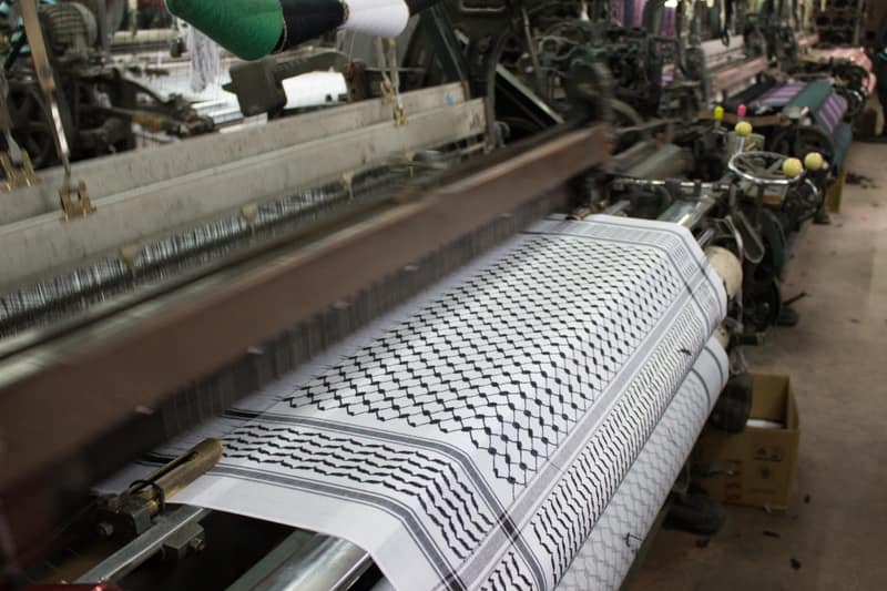 A black-and-white keffiyeh emerging from an industrial loom, the fishnet pattern and its banded border fully formed in the woven cloth. Behind it the machine’s beams, heddles and coloured thread cones recede down the length of the workshop.