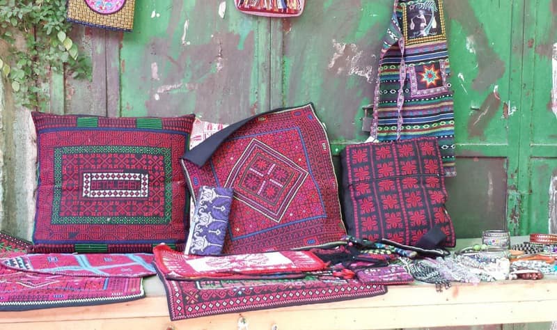 Cross-stitched cushion covers, a long shoulder bag and folded runners propped and laid out on a wooden bench against a chipped green-painted door. The largest cushion is worked in red and green on black with a word cross-stitched across its central panel.