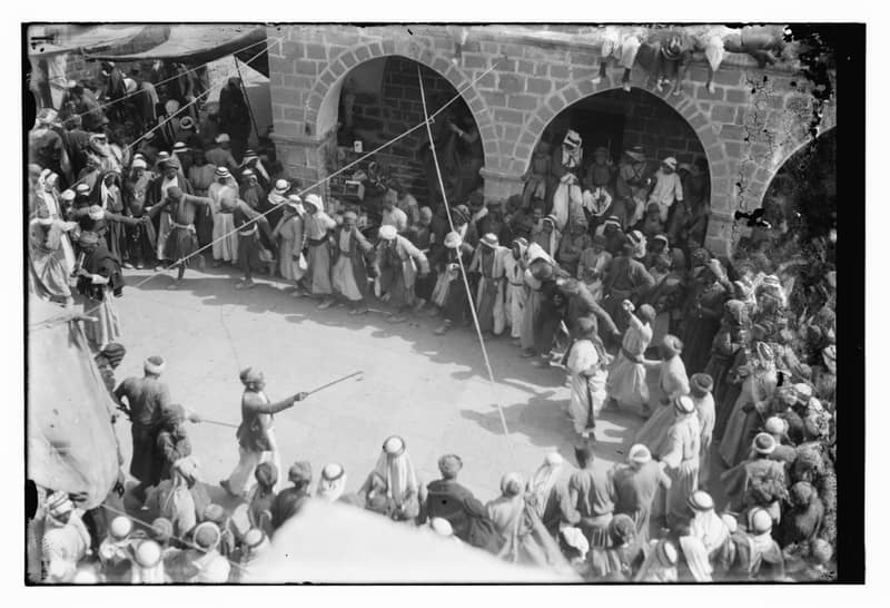 A dry-plate photograph looking down into a stone courtyard. Dozens of men in long robes and white head-cloths move in a long linked line with their arms joined, watched by a dense crowd standing under two arches and packed along the far wall. One man in the foreground holds a cane out at arm’s length.