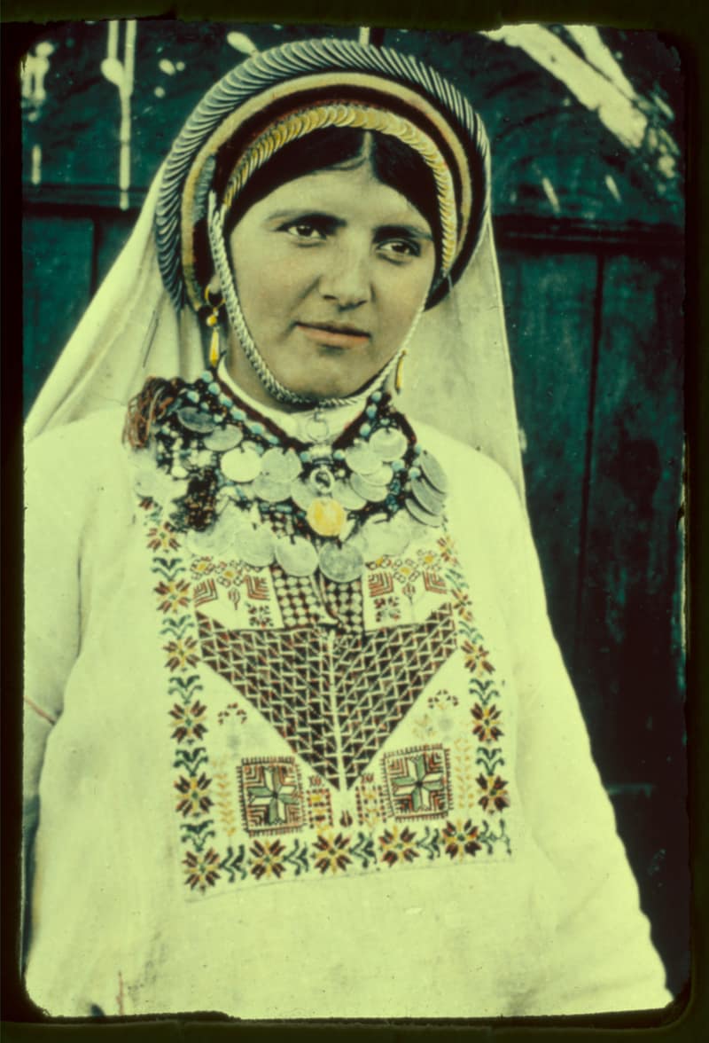 A woman in Ramallah dress photographed in colour. A padded cap ringed with rows of silver coins sits over her parted hair beneath a white veil; a heavy coin necklace lies at her throat, and the chest of her undyed linen dress carries a square panel of red, green and gold cross-stitch.