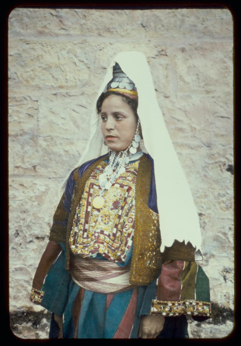A woman in Bethlehem dress stands against a limestone wall. She wears a tall conical cap covered in silver coins under a long white veil, a chain of coins under her chin, a densely embroidered chest panel in red, gold and white, a gold-worked jacket and a striped sash.