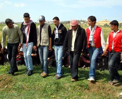 Seven young men in jeans, hoodies and keffiyehs stand shoulder to shoulder in a green field, hands joined in a line, stepping together. Other people sit on the grass behind them; low hills and buildings are visible on the horizon.